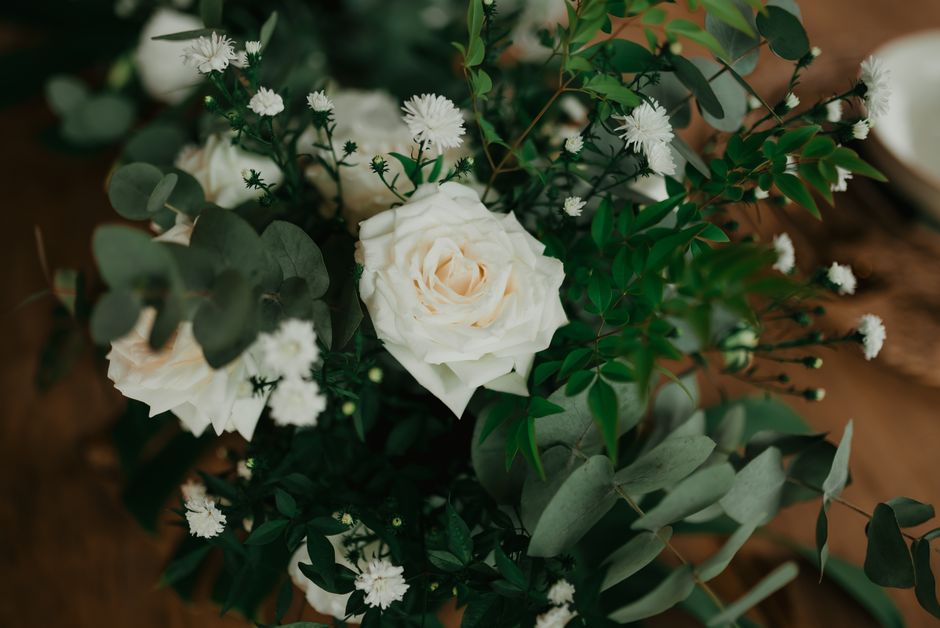 A floral arrangement featuring a central large, fully bloomed white rose with layered petals, complemented by small, white, daisy-like flowers and lush green foliage including eucalyptus leaves. The b