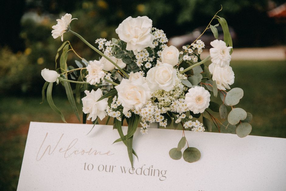 A fresh floral arrangement featuring a bouquet of white roses, delicate white daisies, and small white filler flowers, accented with trailing eucalyptus leaves. The bouquet is set on a white card with