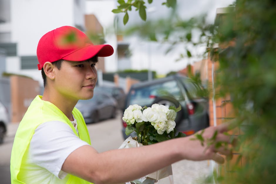 A young male flower delivery courier wearing a red cap and a high-visibility yellow vest is holding a bouquet of white roses and hydrangeas wrapped in white paper. He is reaching out to place the flor