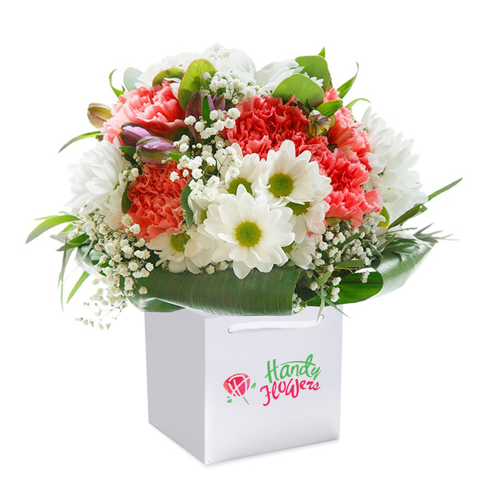 Set against a pure white background, this elegantly composed bouquet, assembled within a square white gift bag, reflects the refined artistry of Surrey's floristry scene. The focus is on a harmonious blend of lush carnation blooms in the softest shades of peach and coral, complemented by radiant red blossoms for dramatic contrast. Their copious, delicately ruffled petals create a plush texture, echoing the layered richness of Surrey's stately gardens. Interspersed throughout, snowy daisies with bright yellow-green centers offer a buoyant charm, reminiscent of meadow wildflowers near Hampton Court or across the Surrey Hills. Sprigs of gossamer baby's breath float among the larger flowers, their gentle white blossoms imparting a sense of airiness and innocence. Nestled subtly to one side, a few closed buds tipped with purple provide visual intrigue, hinting at promise and growth. Surrounding the arrangement, broad, glossy green leaves frame the bouquet, grounding it in a natural, sophisticated structure-much as ancient woodland frames local landscapes. The presentation is completed by a pristine white gift bag with sturdy handles, emphasizing practicality for doorstep gifting across Surrey's market towns. Light falls softly over the bouquet, enhancing its tones and textures, while each stem reflects care and skill. This scene embodies a sense of celebration, tenderness, and locality-ideal for conveying gratitude, love, or well-wishes to family and friends throughout Surrey.